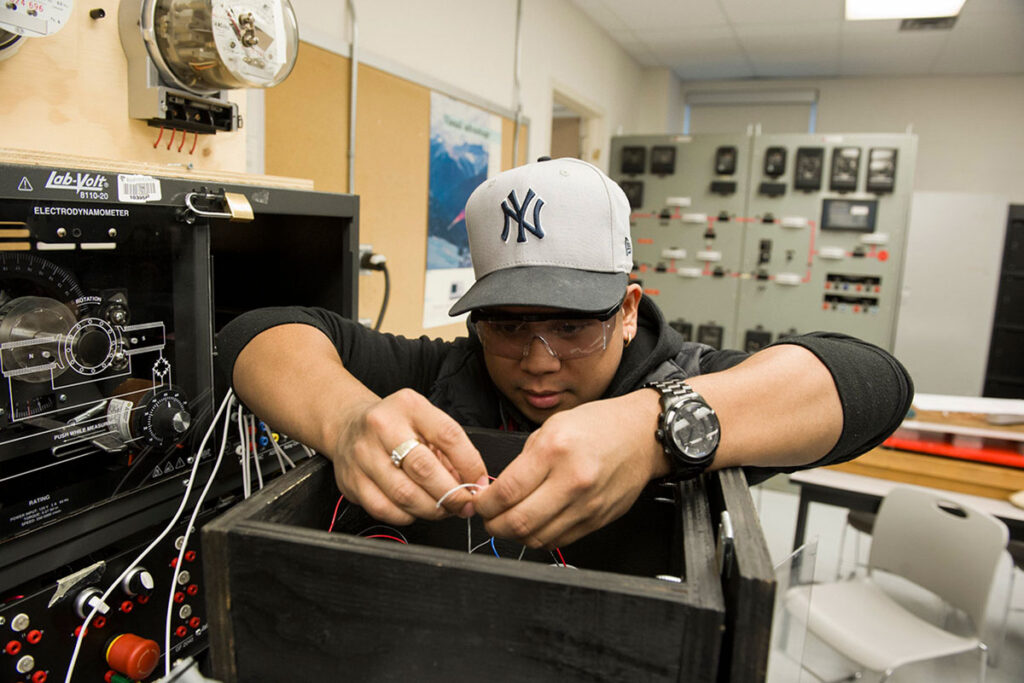 A male student wearing a baseball cap and safety glasses leans over a Lab-Volt electrodynamometer unit, carefully connecting wires inside a wooden enclosure, with industrial electrical panels visible in the background.