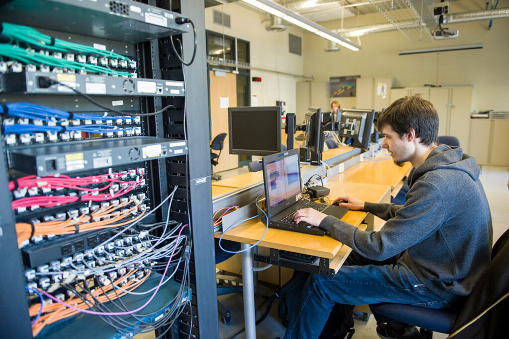 A male student in a grey hoodie types on a laptop at a workstation beside a tall server rack densely packed with multicoloured ethernet cables — green, blue, pink, and orange — in a networking lab.
