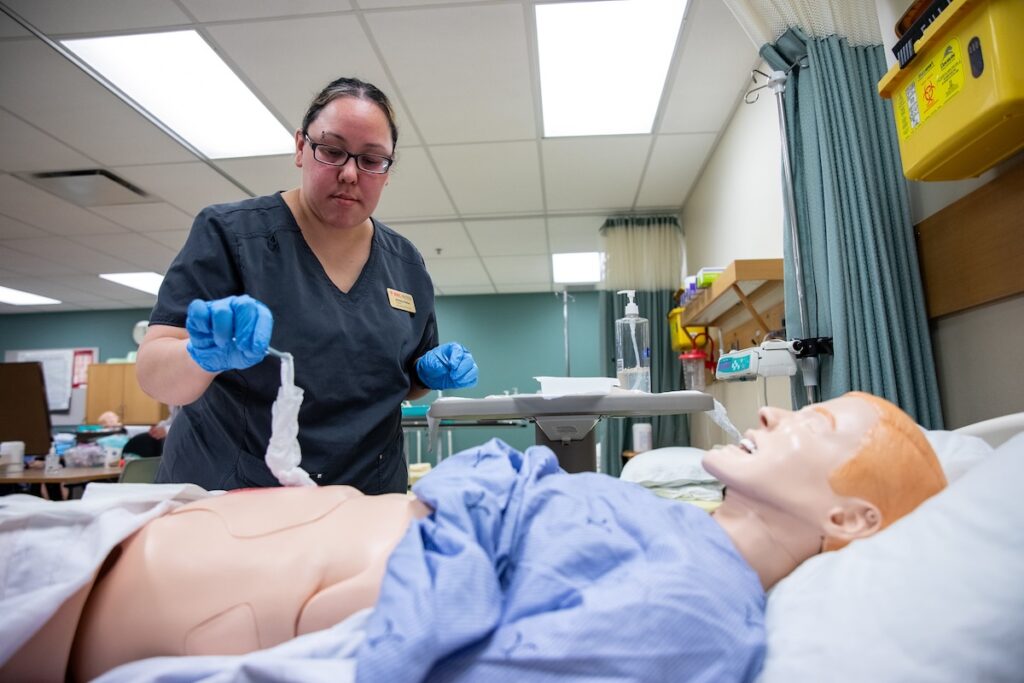 Nursing student practices patient care procedures using a simulation mannequin in a clinical lab.