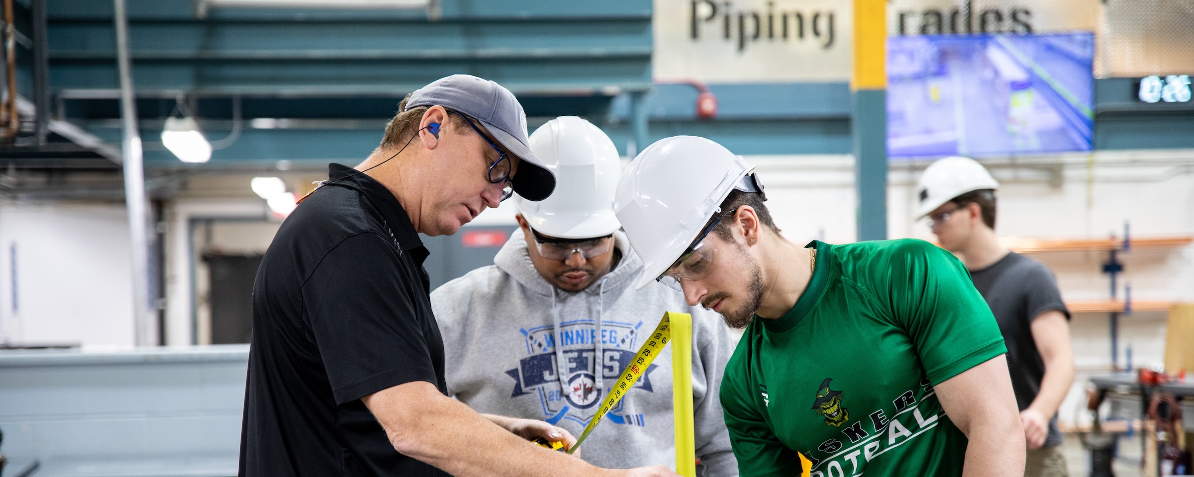 Instructor demonstrates a measurement technique to students wearing hard hats and safety glasses in a piping trades lab.