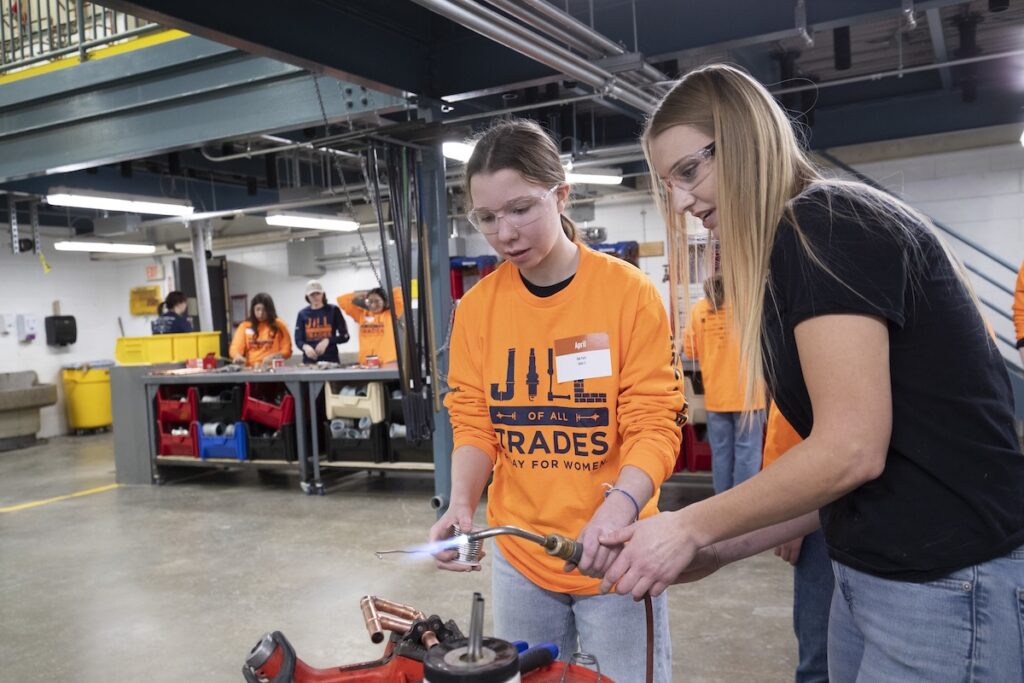 Instructor guides a student using a torch during a hands-on soldering exercise in a piping trades workshop.