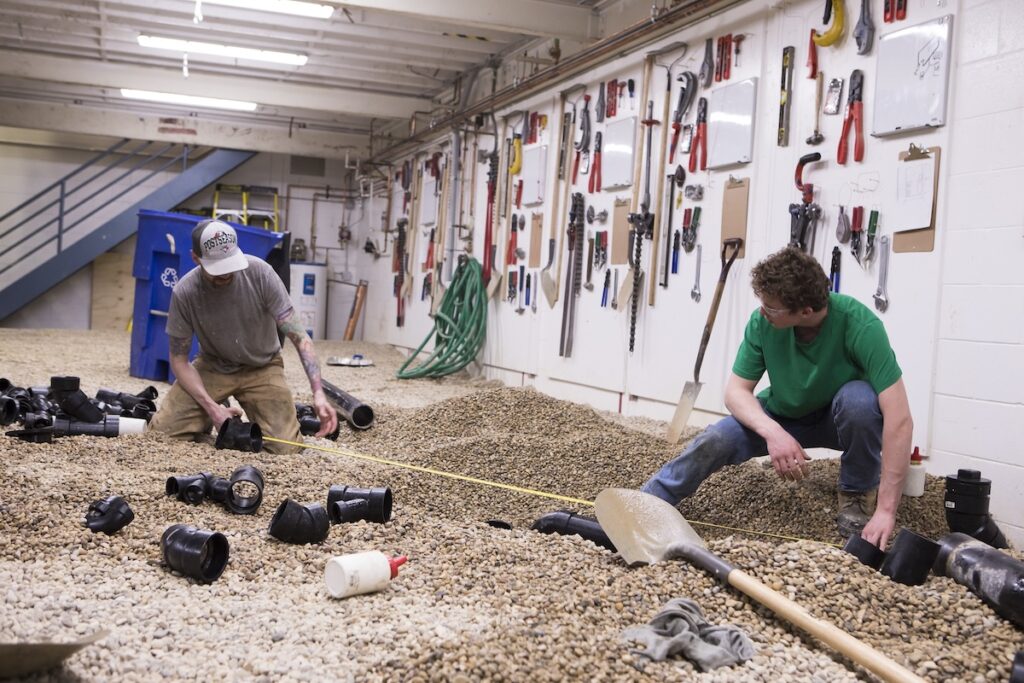 Two piping trades students measure and assemble drainage pipe components during a practical lab.