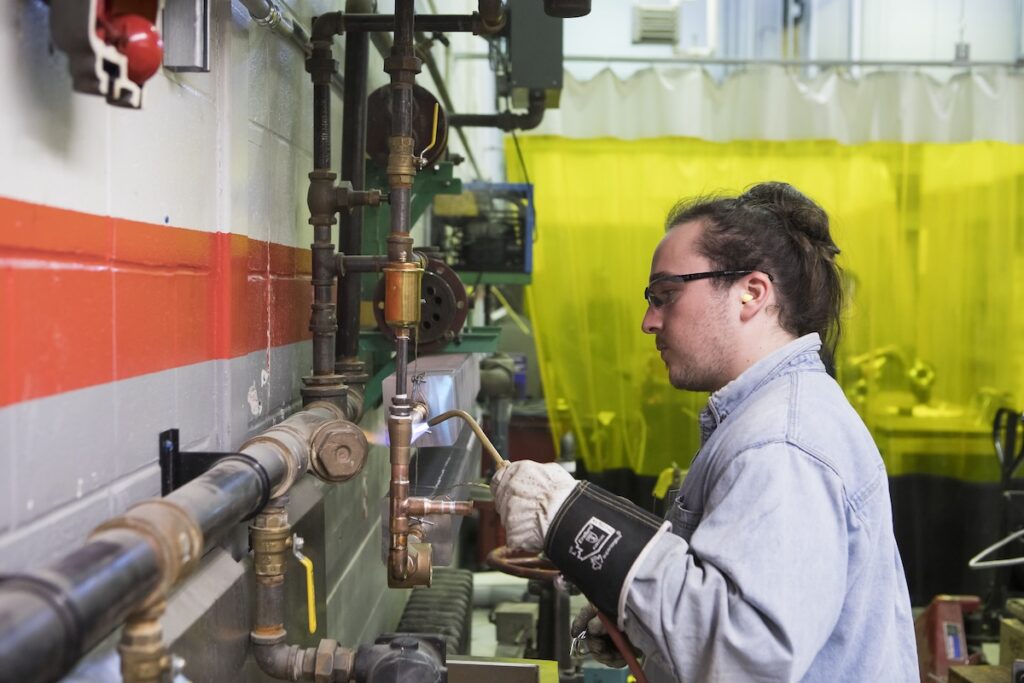 Student brazes copper piping while wearing protective gloves and safety equipment in a trades lab.