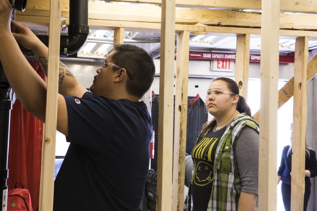 Student installs piping inside a framed wall while another student observes the installation process.