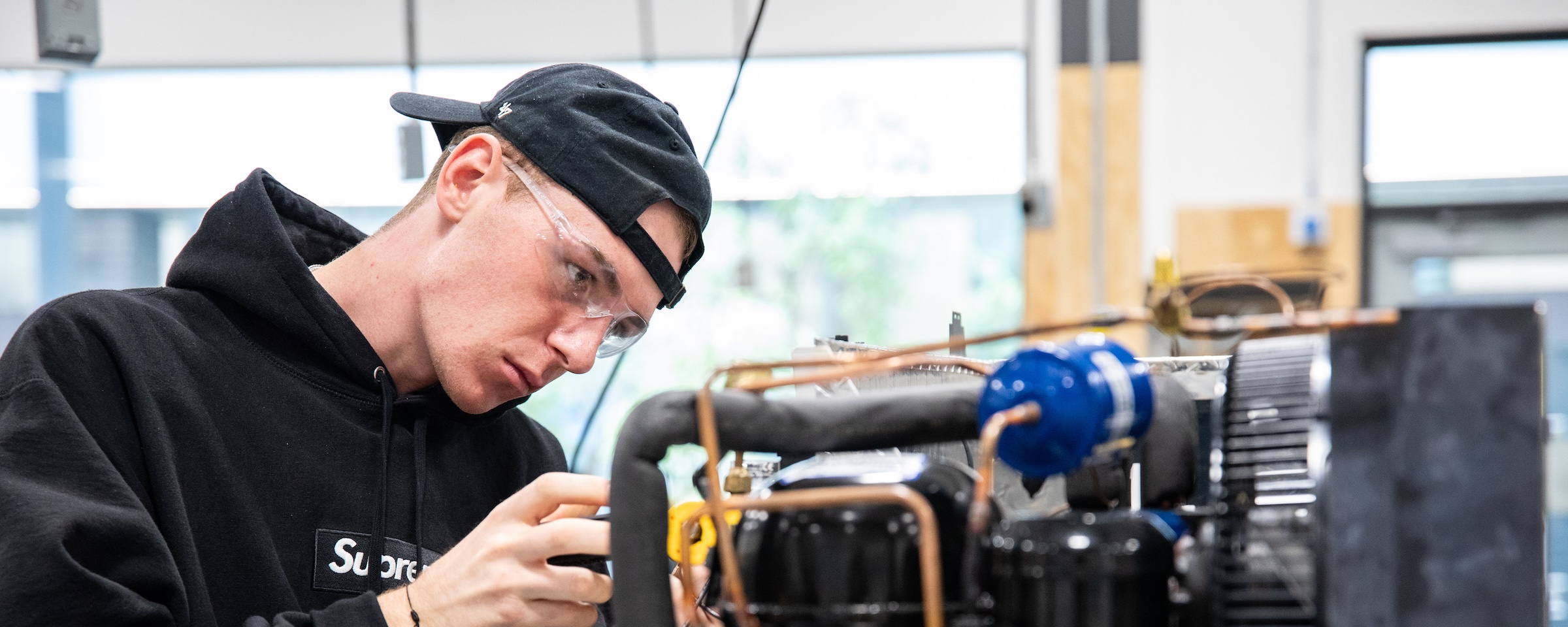 Student wearing safety glasses closely inspects refrigeration equipment and copper piping during a practical lab exercise.