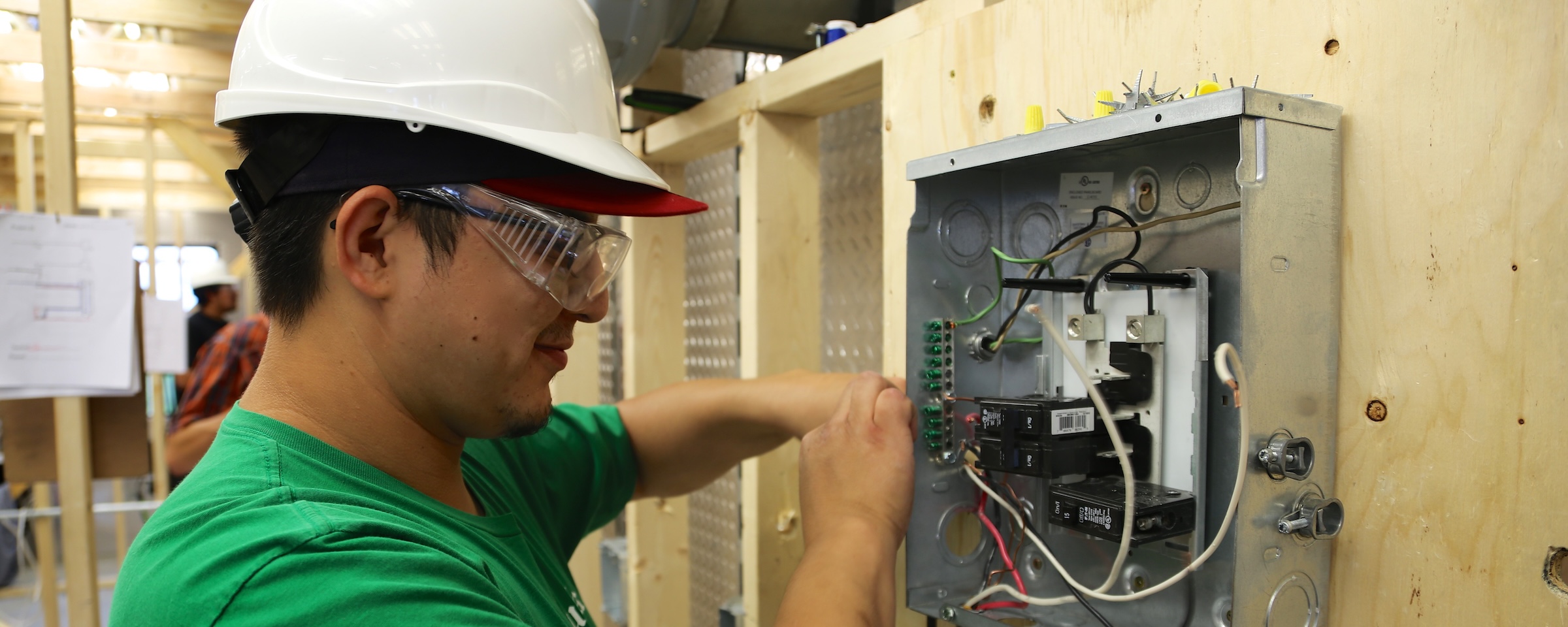 Student wearing safety glasses and a hard hat works on an electrical panel in a trades lab.