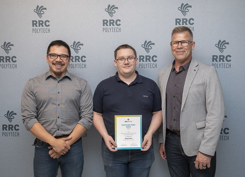 Three people stand in front of a grey background while smiling.