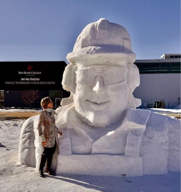 A woman stands next to a snow sculpture of a person's head with a hard hat and goggles.