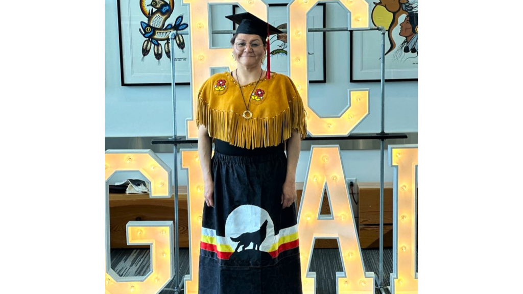 a person in a grad cap and ribbon skirt stands in front of RC Grad lights and Indigenous artwork