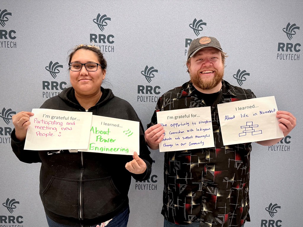 Two people stand beside and hold up signs, while smiling at the camera.