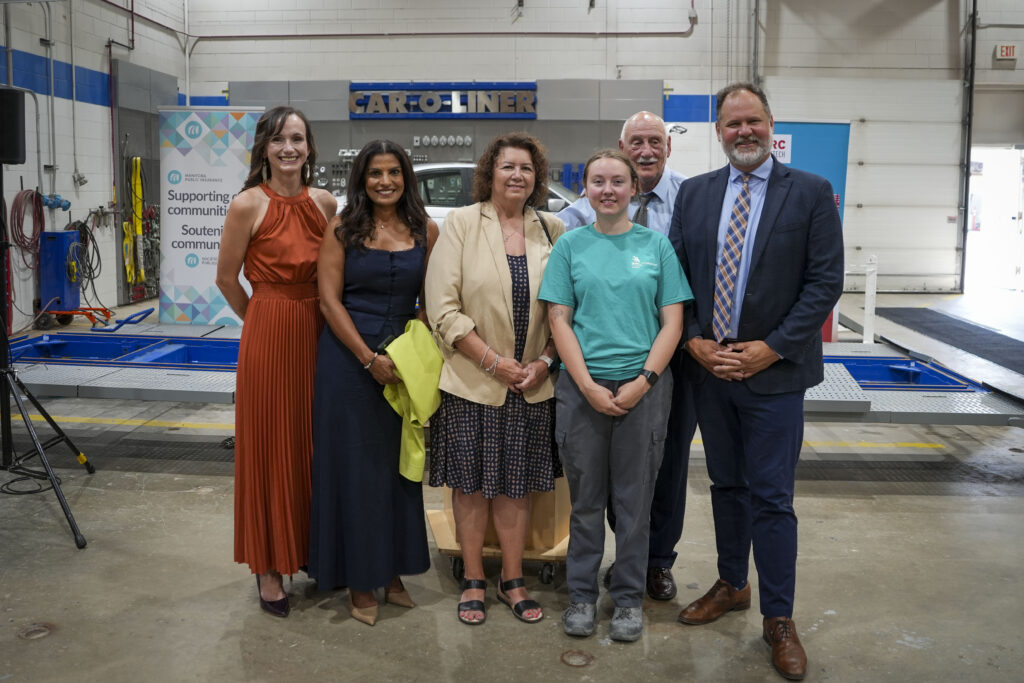 A group of people stand in an autobody shop, and smile at the camera.