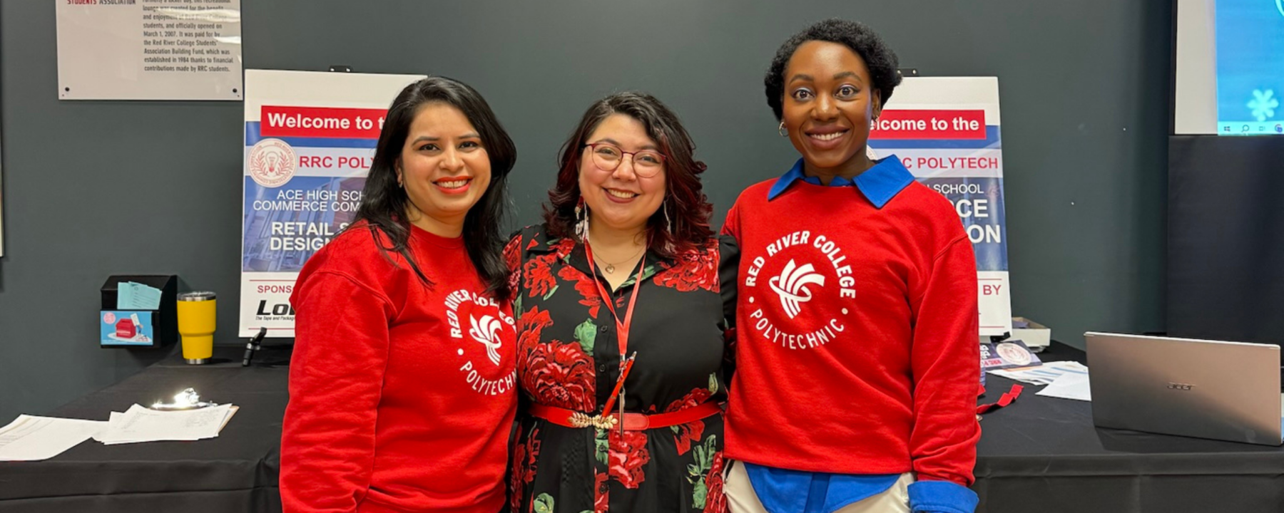 Three female Teacher Education students stand shoulder to shoulder. Two of them wear bright red sweaters, the one in the middle wears a black dress with red flowers.