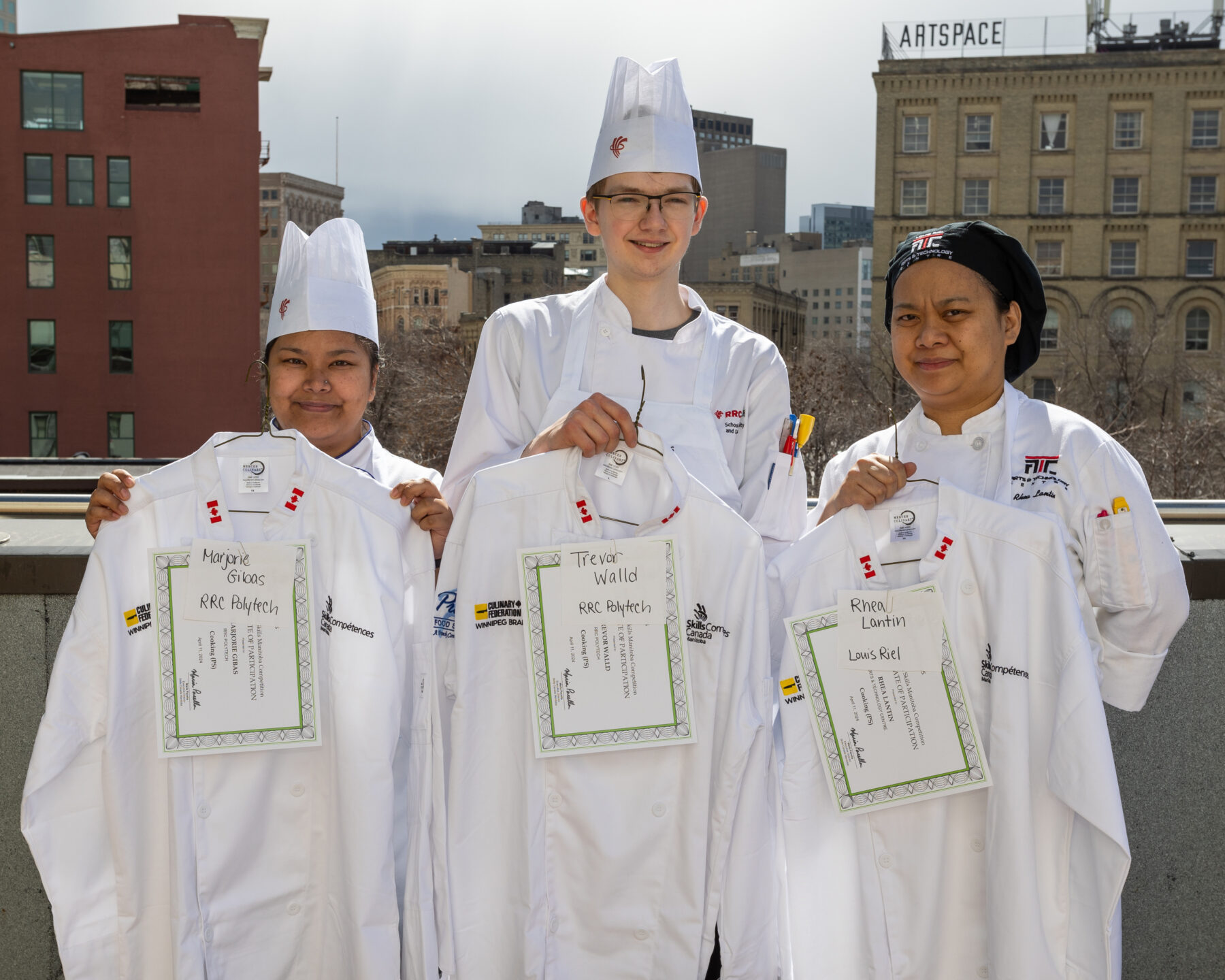 Three culinary students stand in the chef's white atop a rooftop parkade. They are holding winners certificates in front of them and smiling.