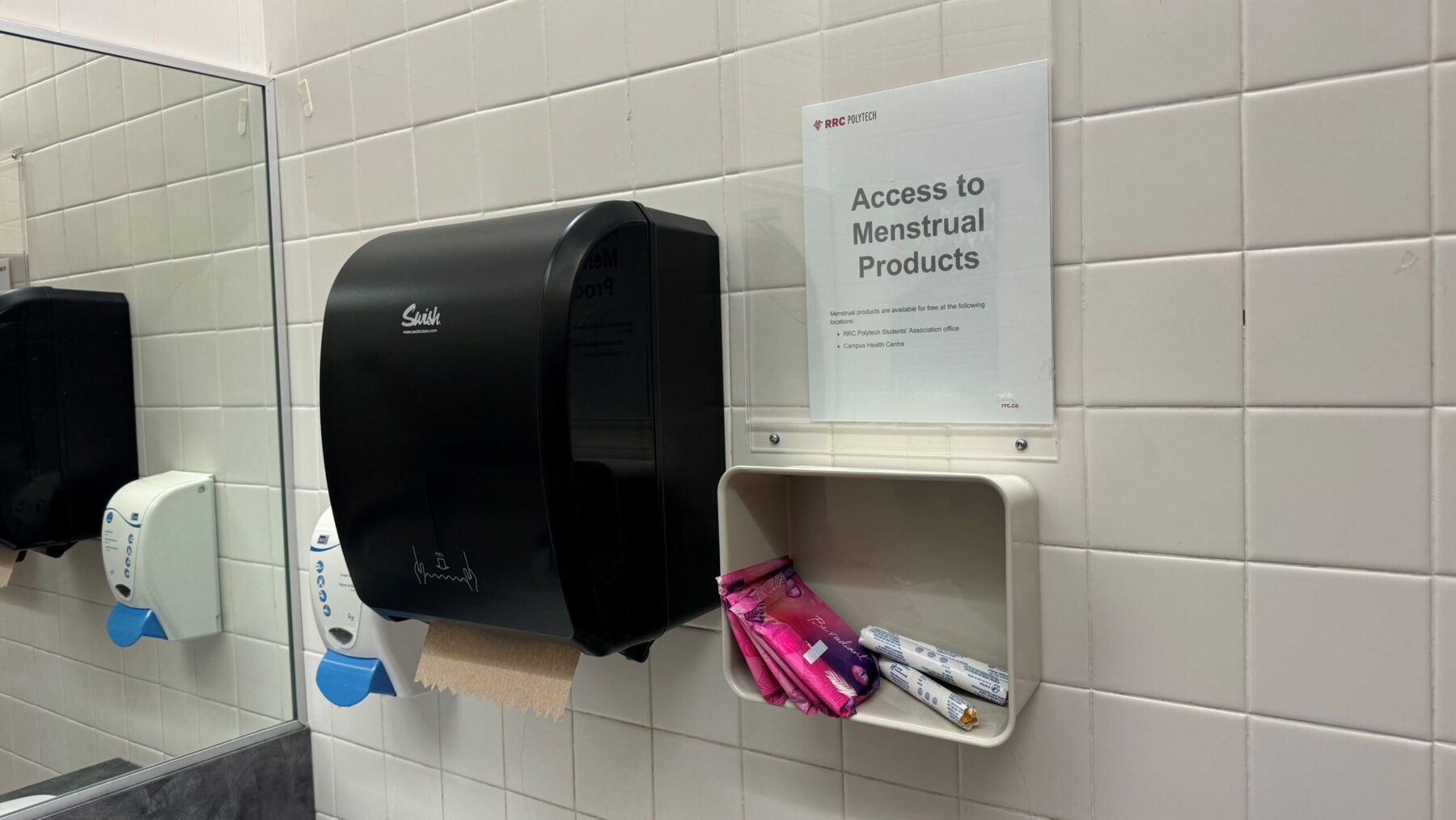 A basket of menstural products on a white tile bathroom wall. A black paper towel dispenser and white soap dispenser are on the wall beside it.