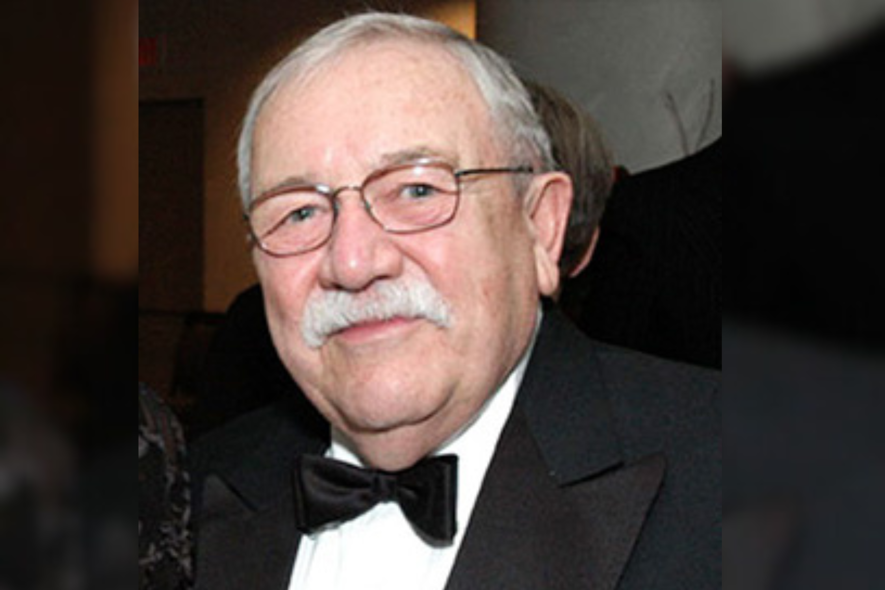 A man wears a black tuxedo jacket and bowtie with a white button up shirt. He has grey hair and metal framed glasses. He looks forward and smiles lightly.