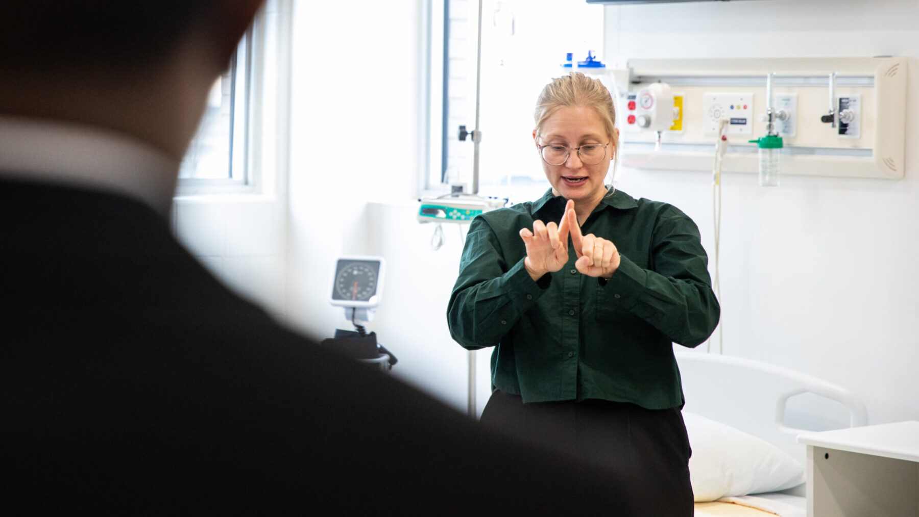 Woman doing ASL in a healthcare facility. Her hands are raised in front of her. There is a man blurred out in the foreground.