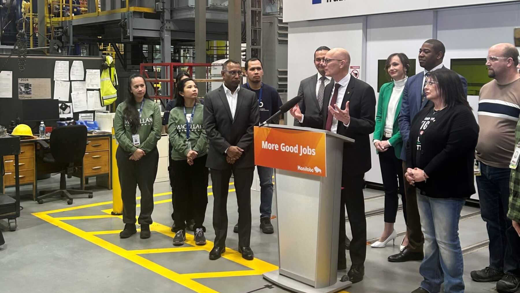 Fred Meier, stands in a warehouse style room at a podium addressing a crowd that is not in view. He is flanked by a group of men and women including employees from Magellan and members of the Manitoba Government.
