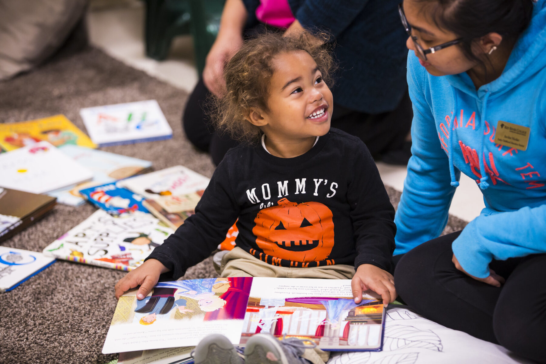 A child sits on the ground while holding a picture book, she is looking at a caregiver and smiling.