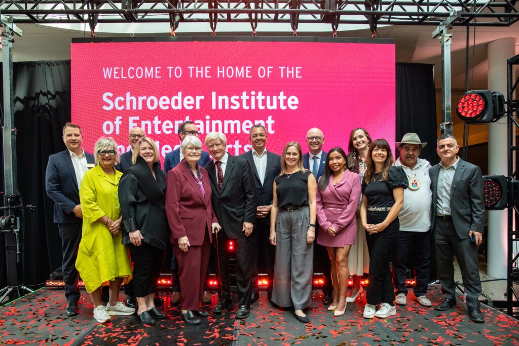 A group of people stand on a stage in front of a large red banner that says, "Welcome to the Home of the Schroeder Institute of Entertainment and Media Arts."