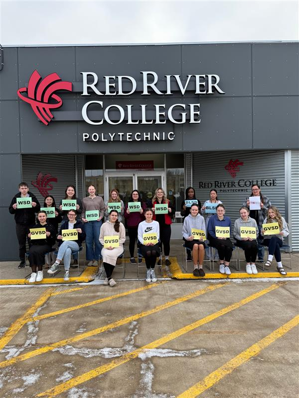 A group of people stand in front of a building with a Red River College Polytechnic sign on the front. They hold signs with the names of their school divisions and smile at the camera.