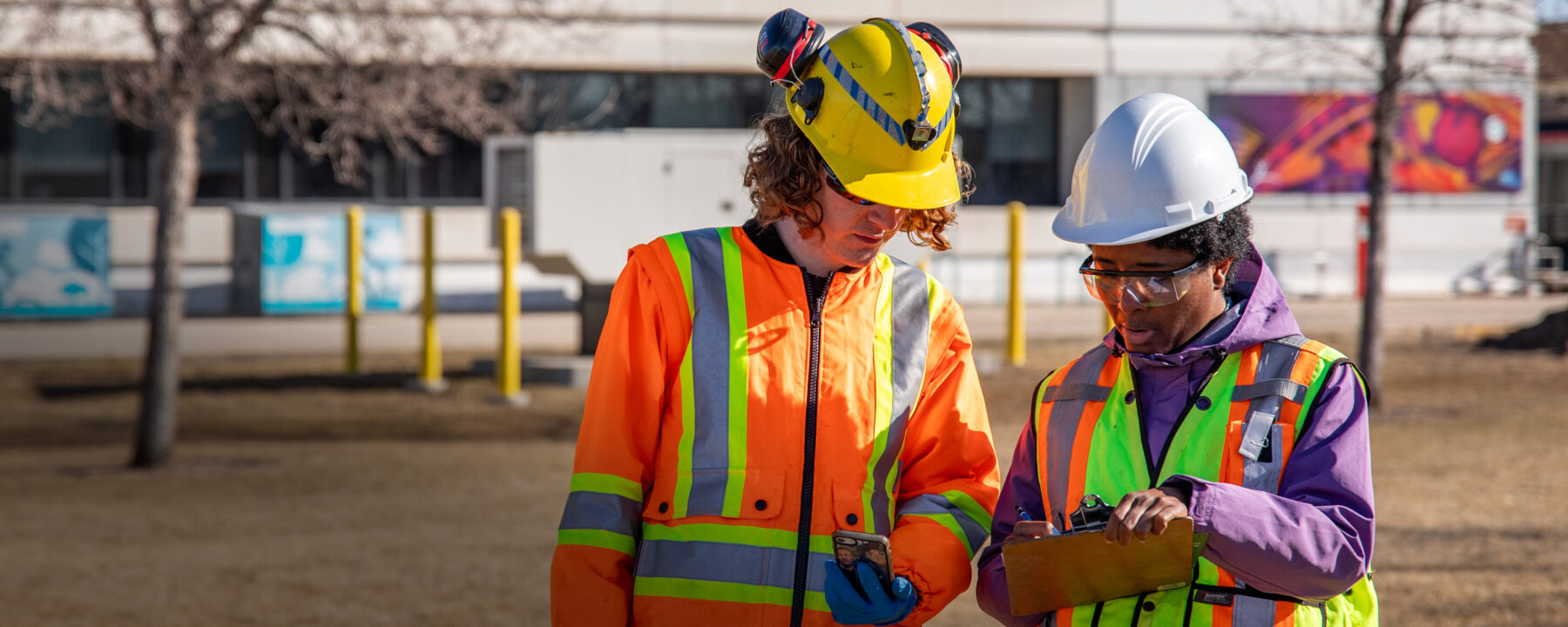 Two people in personal protective equipment stand outdoors and consult a clipboard.
