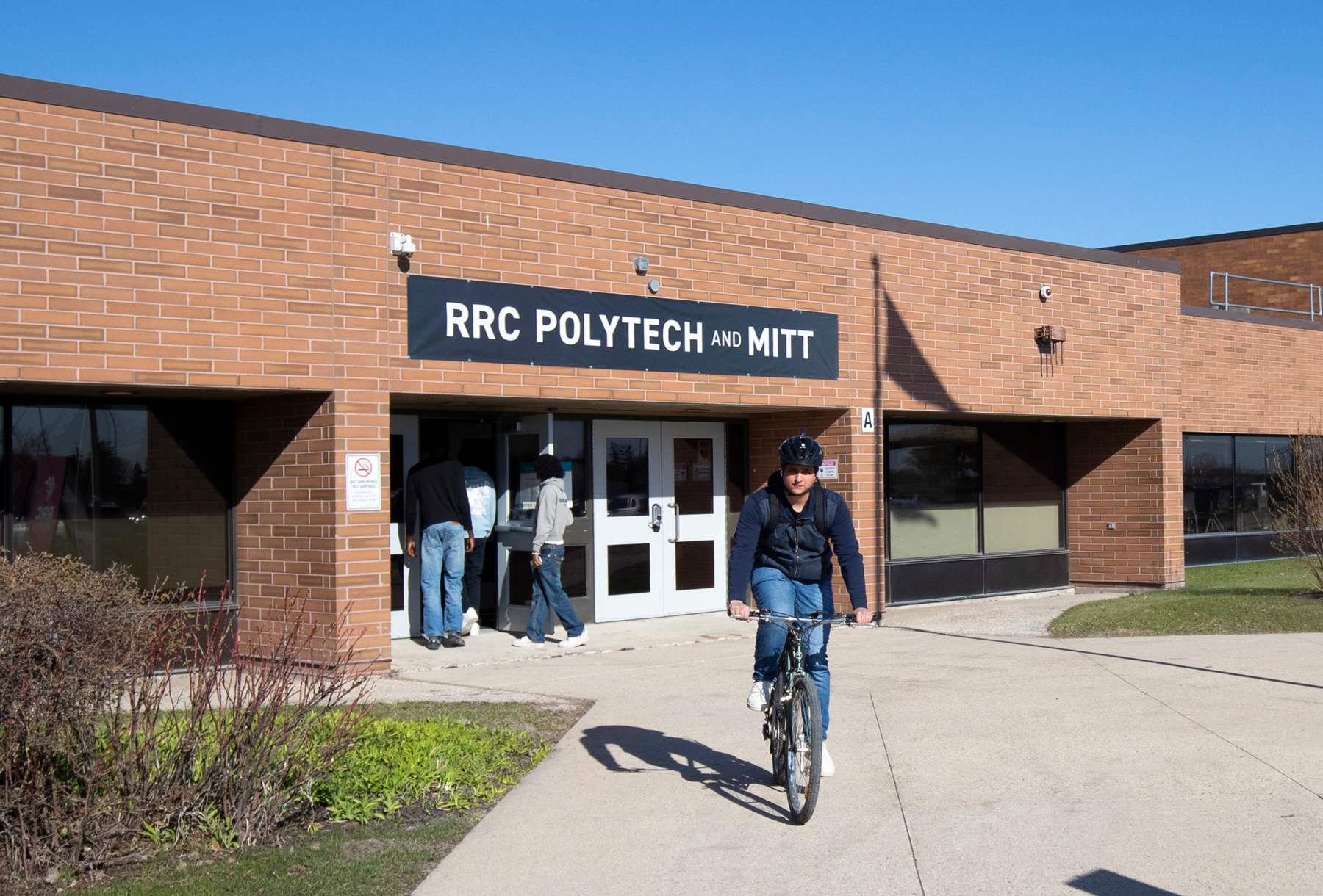 Person riding a bicycle past the entrance of a brick campus building, while two people enter through the front doors beneath an RRC Polytech and MITT banner.