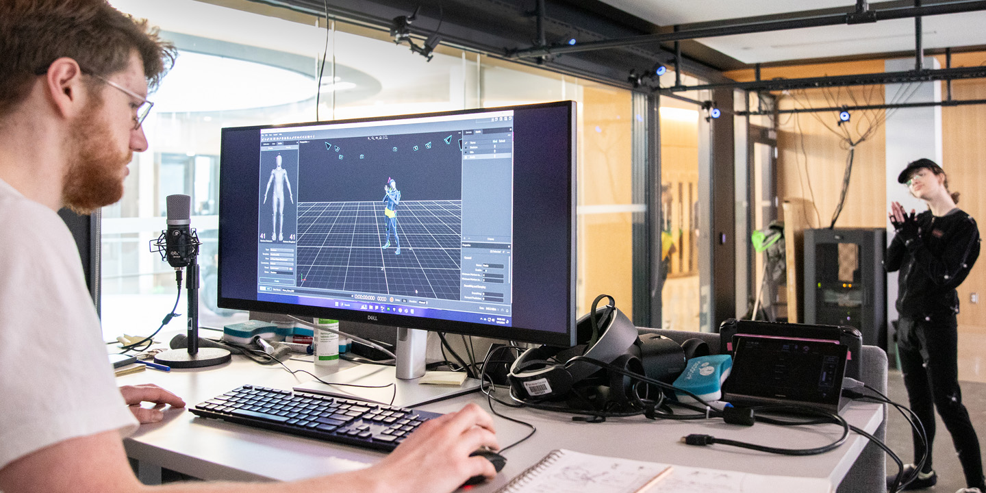 A person works at a screen while another person poses in front of it.