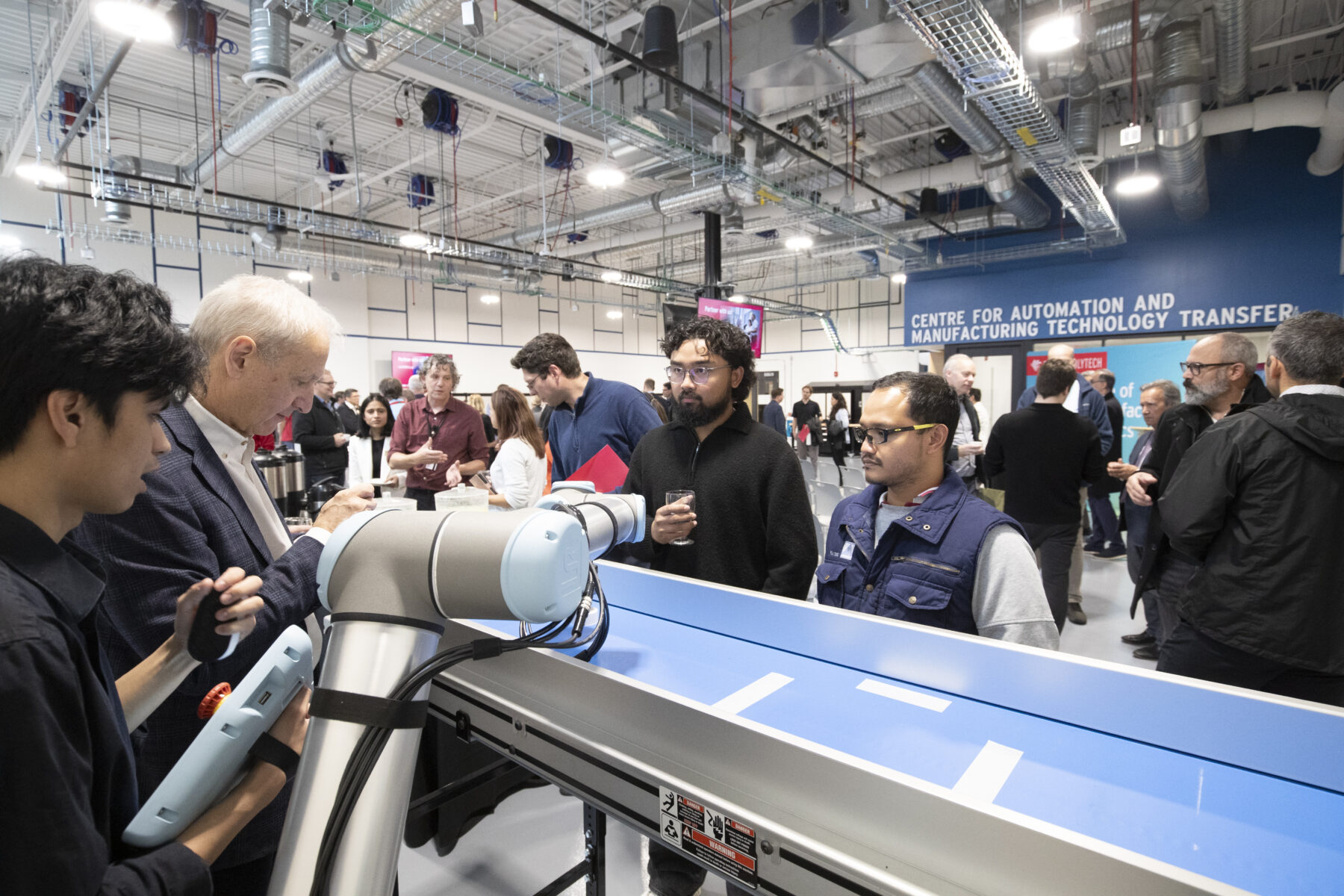 A room filled with people, some people are holding glasses or small plates of food to indicate it is a reception event, the words 'Centre for Automation and Manufacturing Technology Transfer' are on a wall. Some people are looking at equipment demonstrations.