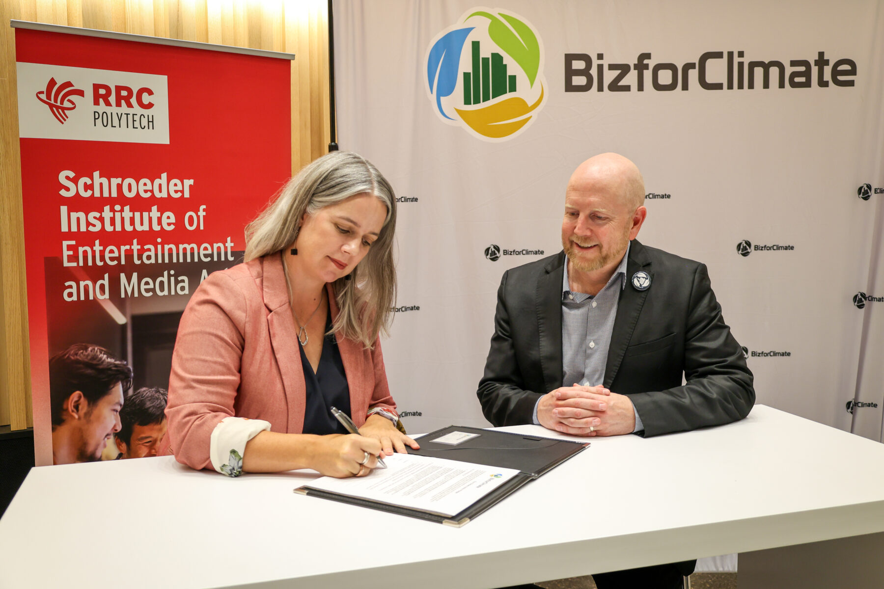 Two people at a desk, with the person on the left signing a document. In the background there are banners for RRC Polytech and BizforClimate.