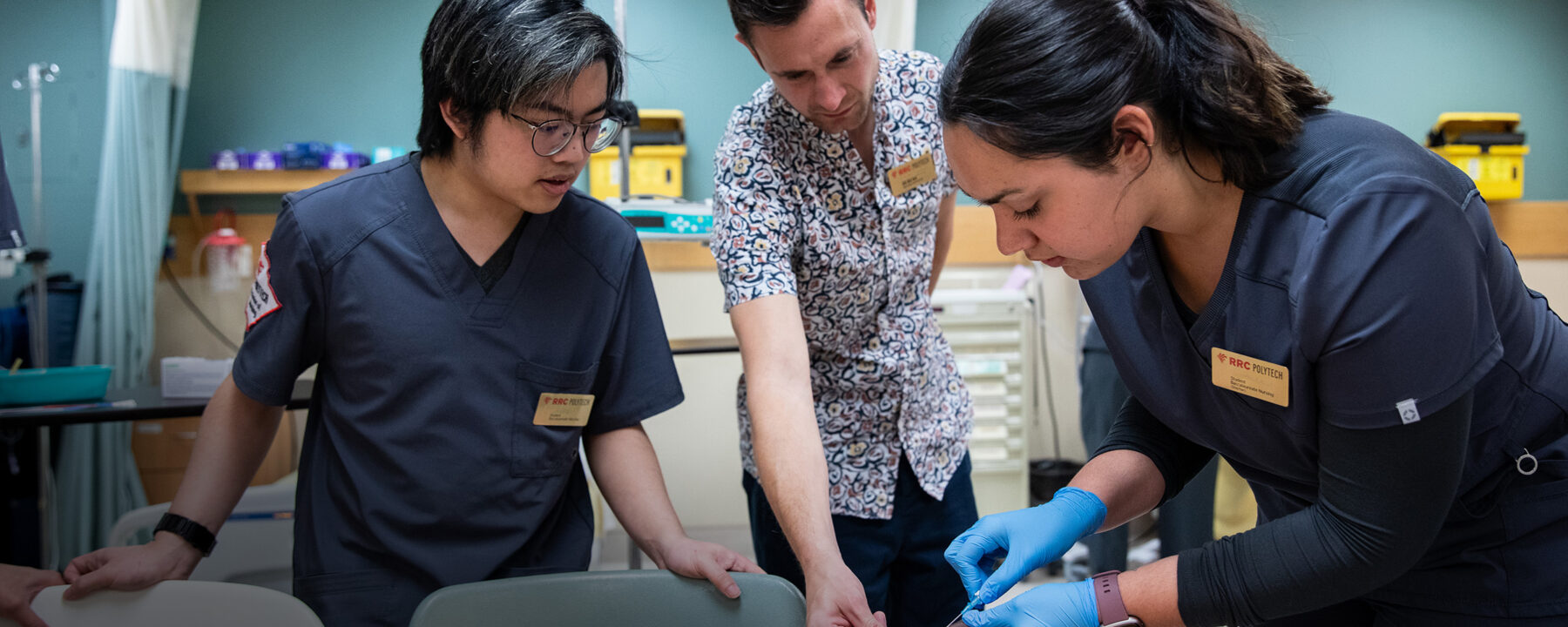 Two nursing students standing on either side of a nursing instructor, while the student on the right wearing blue medical gloves inserts a needle into a mannequin arm.