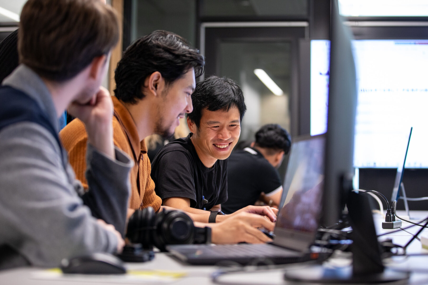 Two people smiling while working on a computer together in a classroom. In the foreground, a person sits with their chin in their fist.