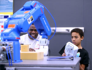 Young boy operates robotic arm under instructor's supervision at manufacturing camp for youth.