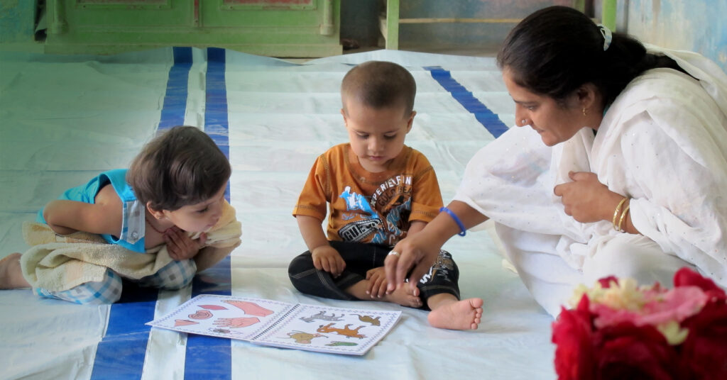 Pakistani woman seated on blanket, looking at picture book with nursery-aged children