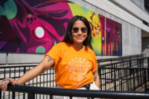 RRC grad Leticia Spence, wearing sunglasses, standing in front of Indigenous mural at Notre Dame Campus.