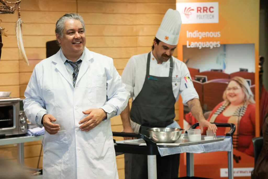 Two RRC instructors — one in lab coat, one in chef's uniform — demonstrating how to cook bannock.