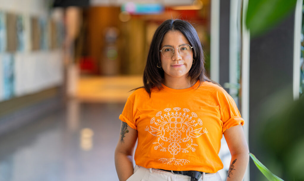 RRC Polytech grad Leticia Spence, in shirt she designed for Orange Shirt Day 2023, standing inside at the Notre Dame Campus.