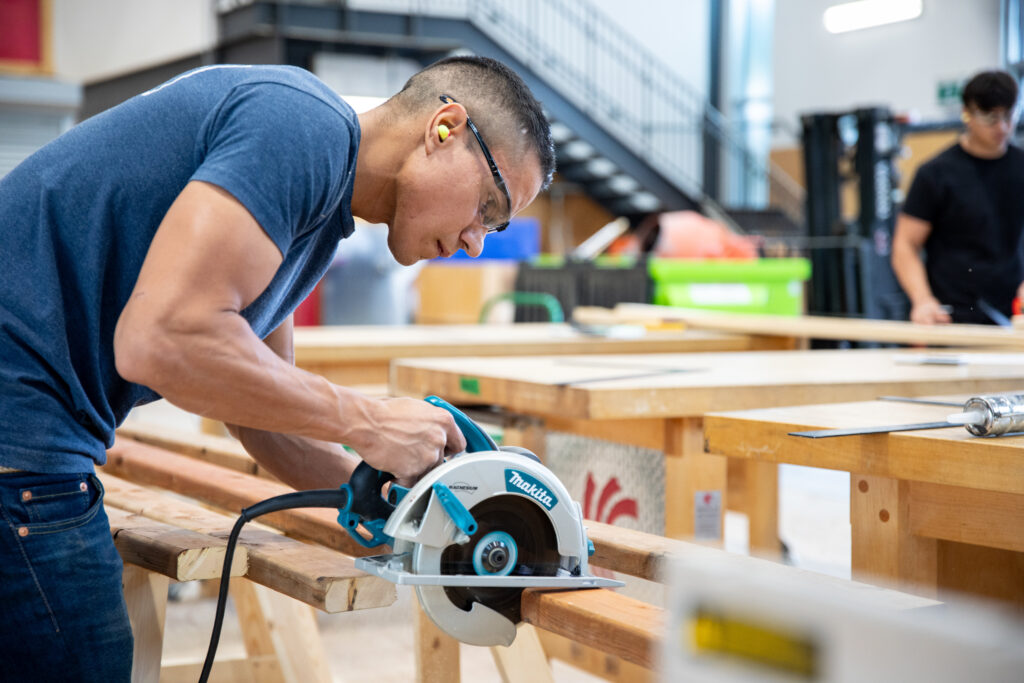 Male student, wearing earplugs and protective eyewear, using circular saw to cut wood inside RRC Polytech carpentry lab.