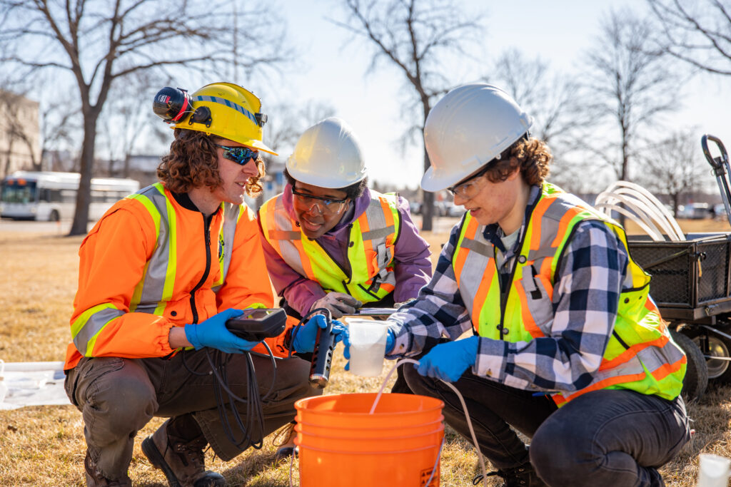 Trio of engineering students wearing safety vests, kneeling around a pail while testing water quality outdoors
