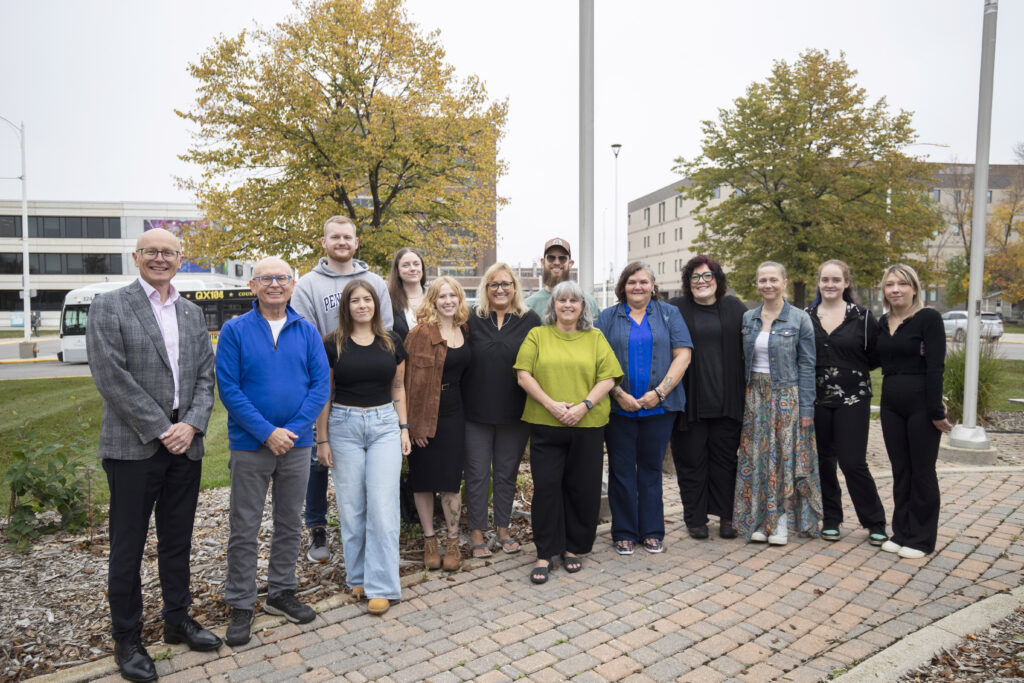 A group of people, including students and instructors from RRC Polytech's ASL-English Interpretation program, standing in a line outside the Notre Dame Campus flagpoles.