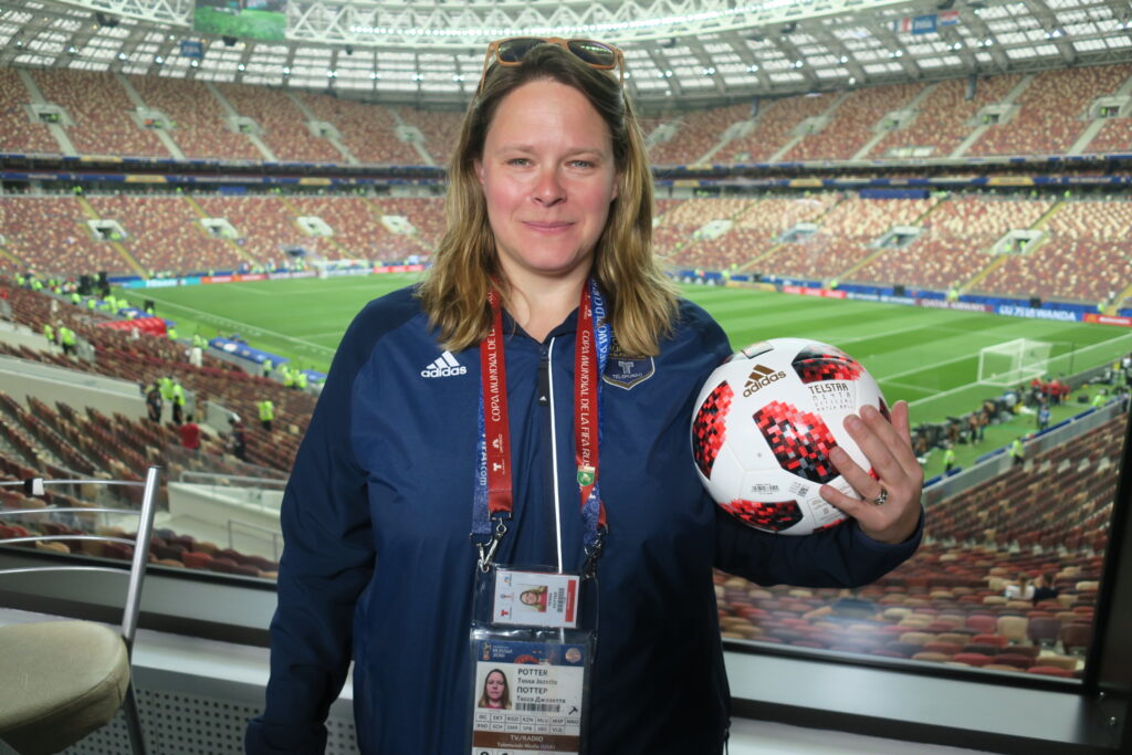 Shot of Tessa Potter, wearing press credentials and holding soccer ball, standing in the press box of a FIFA World Cup stadium in Russia.