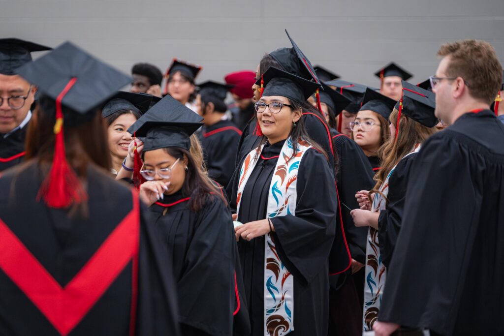Female student wearing new Indigenous stole, surrounded by other graduates in caps and gowns.