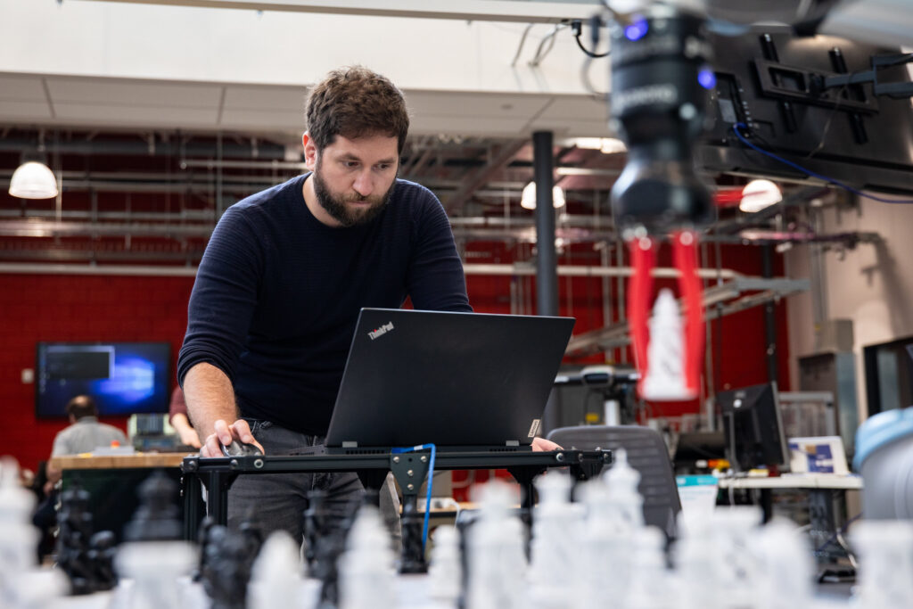 Male RRC Polytech researcher works controls to move a robotic arm clasping a chess piece.