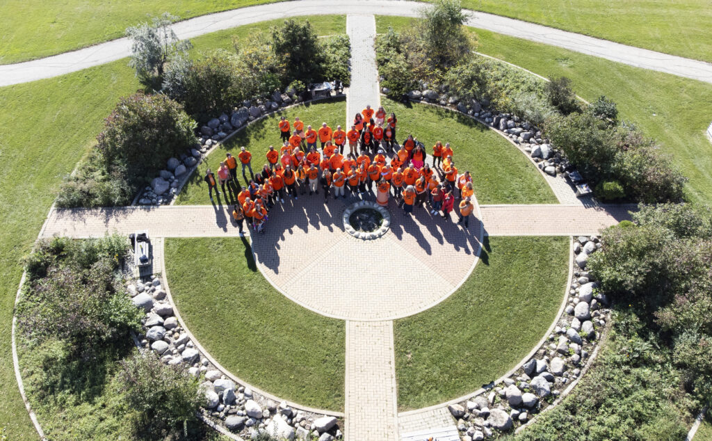 Aerial shot of RRC Polytech staff and students, all wearing orange shirts, gathered outside in the Medicine Wheel garden.