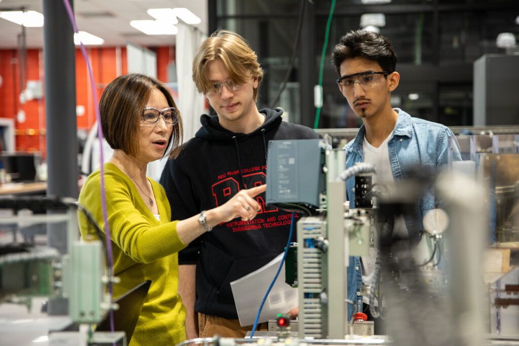 Female instructor programs mechatronics equipment as two male students look on.
