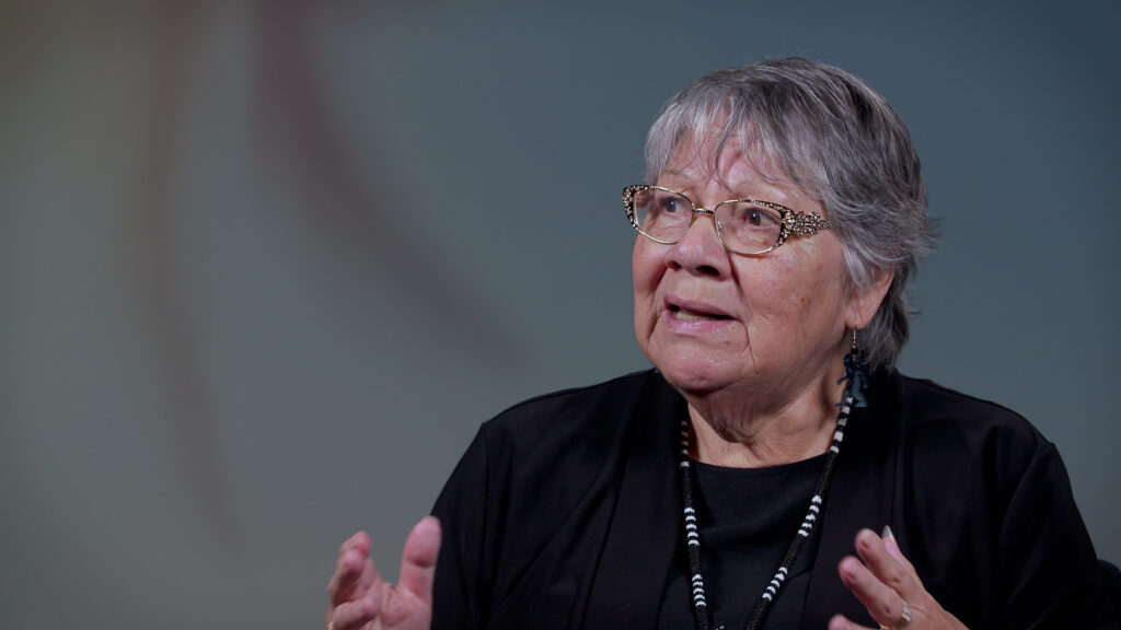 Older Indigenous woman, wearing glasses, black top and beaded necklace, being interviewed.