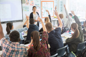 Teacher standing at the front of a classroom with students raising their hands