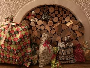 Gifts wrapped in festive fabric gift bags in front of a fireplace