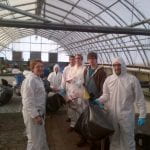 A group of students in protective gear hold up trash bags in a greenhouse.