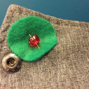 Green felt circle with a red bead pinned to a grey jacket in front of a blue background
