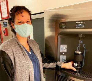 A woman wearing a mask filling a reusable mug at a water bottle fill station
